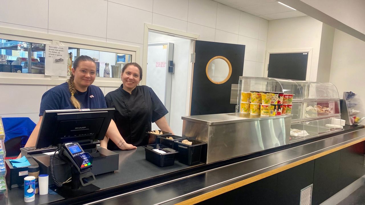 To smilande medarbeidarar, Rebekka og Yulia i kantina ved Laksevåg og Bergen Maritime vgs, står klare bak disken for å servera elevane. Den eine er kledd i ei mørkeblå T-skjorte og den andre i ei svart kokkejakke. Kantina verkar rein og ryddig, med eit utval av matvarer som nudlar og wrapar synleg for sal. Ved lunsjtider blir det også servert varm mat etter ein variert meny. Ved kassa står ein betalingsterminal klar til bruk, og atmosfæren verkar imøtekommande og triveleg